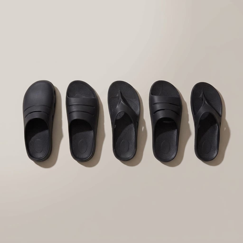 Five Fanture styles in black laid side by side from above: a clog, two slides and two flip flops