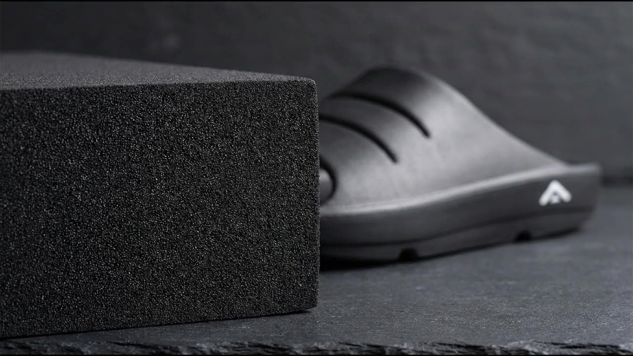 A slow pass across a sliced block of solid black closed-cell foam, its fine even cells catching the light, toward a black Fanture recovery slide behind it on dark slate