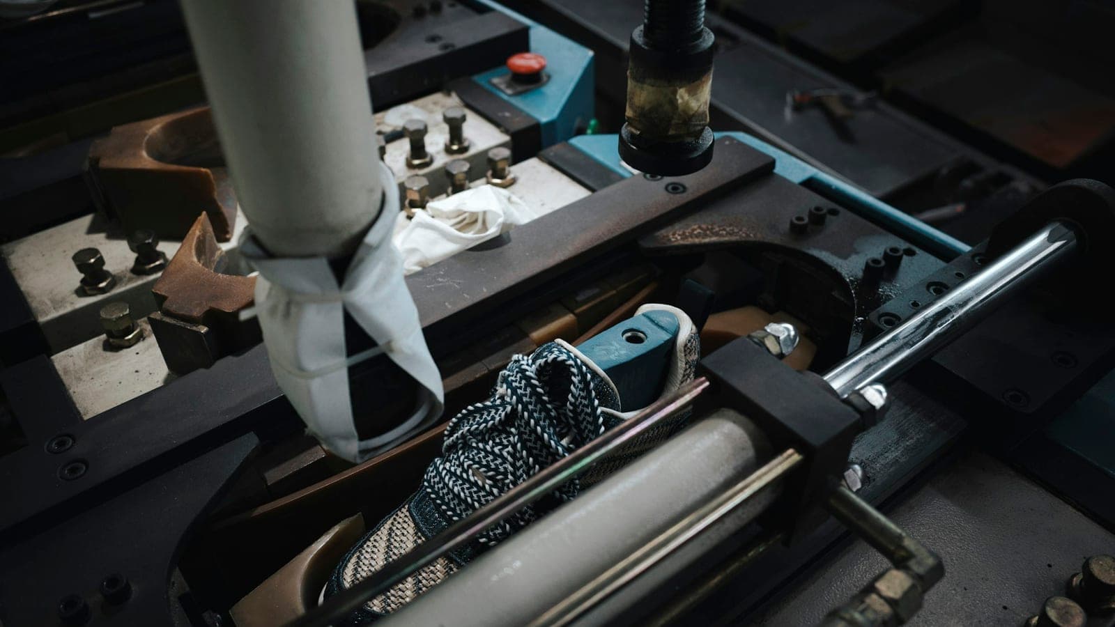 A shoe held inside industrial footwear machinery on a factory assembly line, the metal tooling closed around the sole