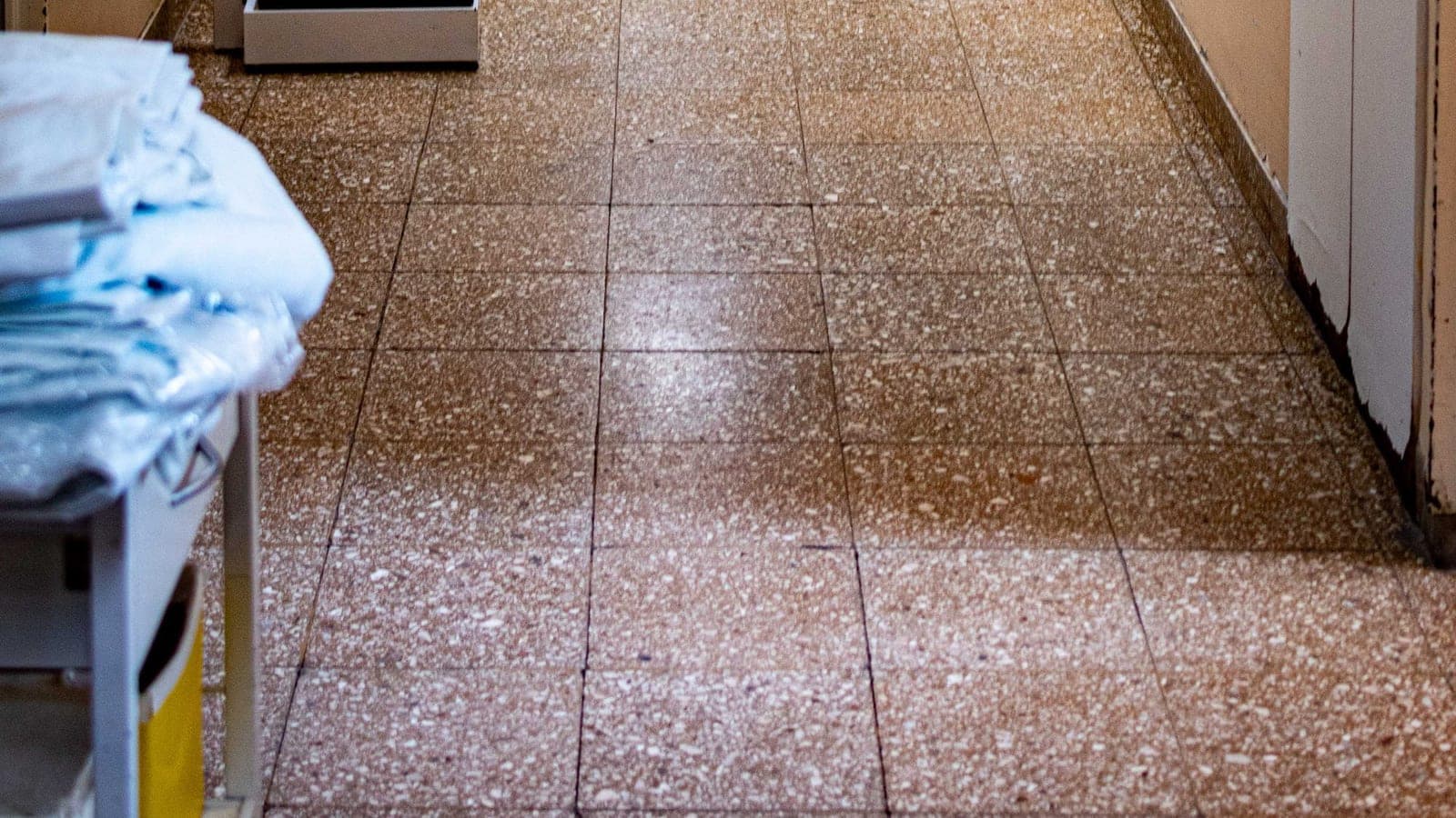 A hard speckled terrazzo floor running down a hospital corridor, a linen trolley at the edge of the frame