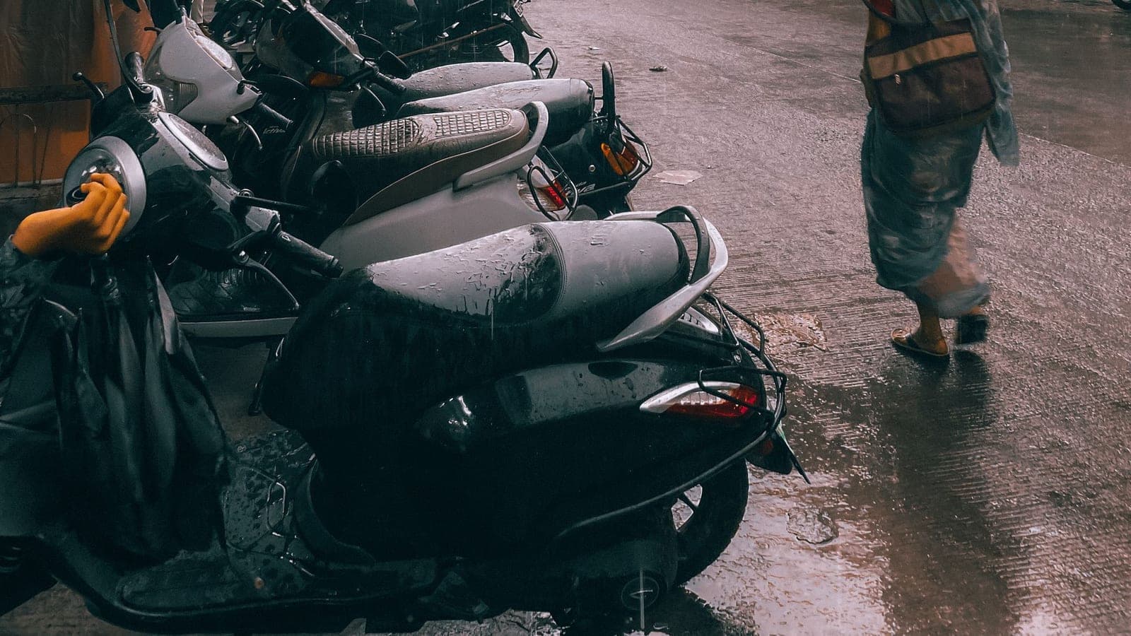 A row of scooters parked in heavy monsoon rain, water running across the road, someone walking past through it in open sandals