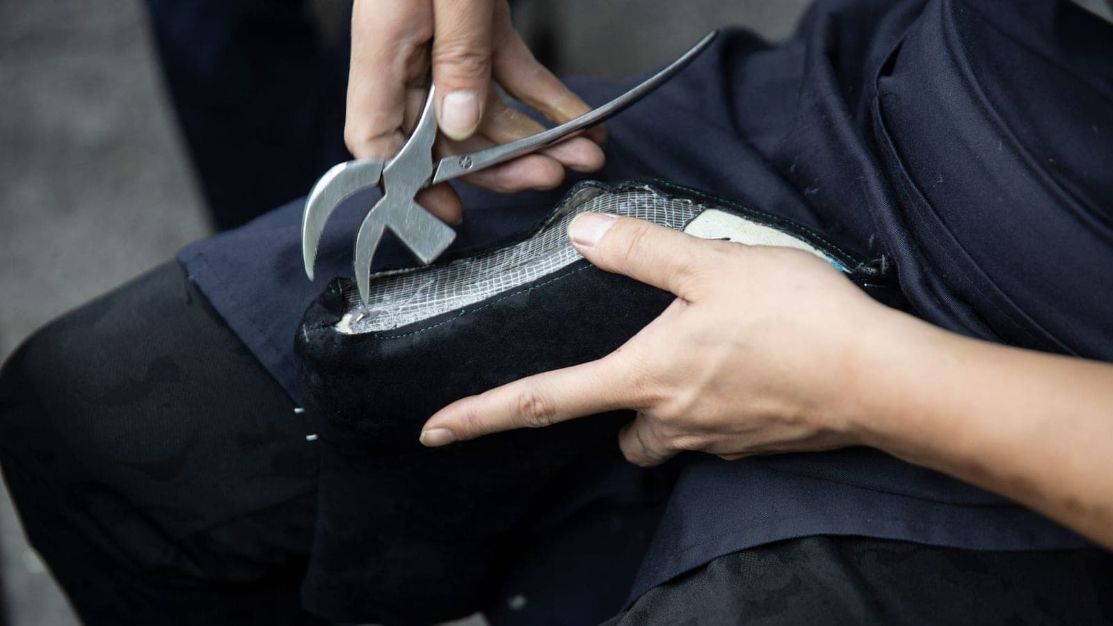 A cobbler's hands close up at a workbench, working the edge of a leather shoe with a pair of pliers
